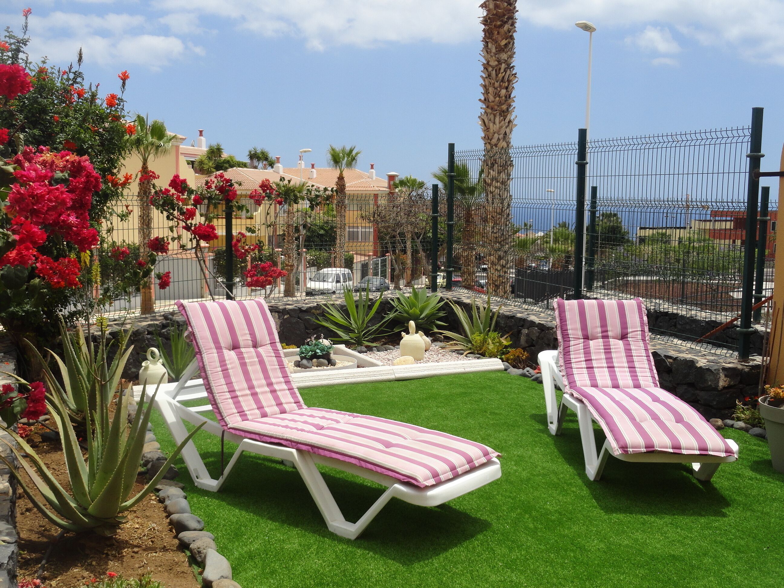 Deux fauteuils de jardin blancs avec des coussins à rayures roses, un petit jardin avec des fleurs rouges et des plantes vertes, et une vue sur une zone côtière avec des bâtiments et une clôture en arrière-plan.