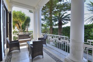 A covered outdoor seating area with wicker furniture, white columns, and a view of tropical trees.