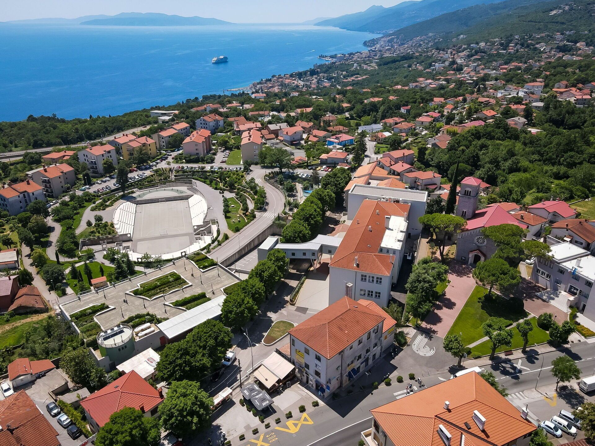 An aerial view of a coastal town with a prominent church, a large open plaza, and residential buildings with red-tiled roofs.