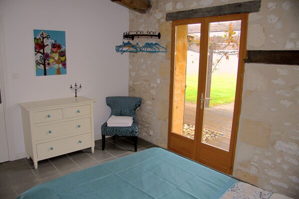 Una habitación con una cama, una silla, un aparador, una puerta que da al exterior y un adorno colgado en la pared.