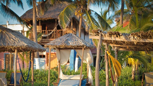 Un lit installé sur la plage, surmonté d’un toit de chaume, entouré de palmiers et d’une structure en bois.