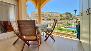 Un balcón con sillas de madera y una mesita, con vista a una piscina y edificios residenciales.