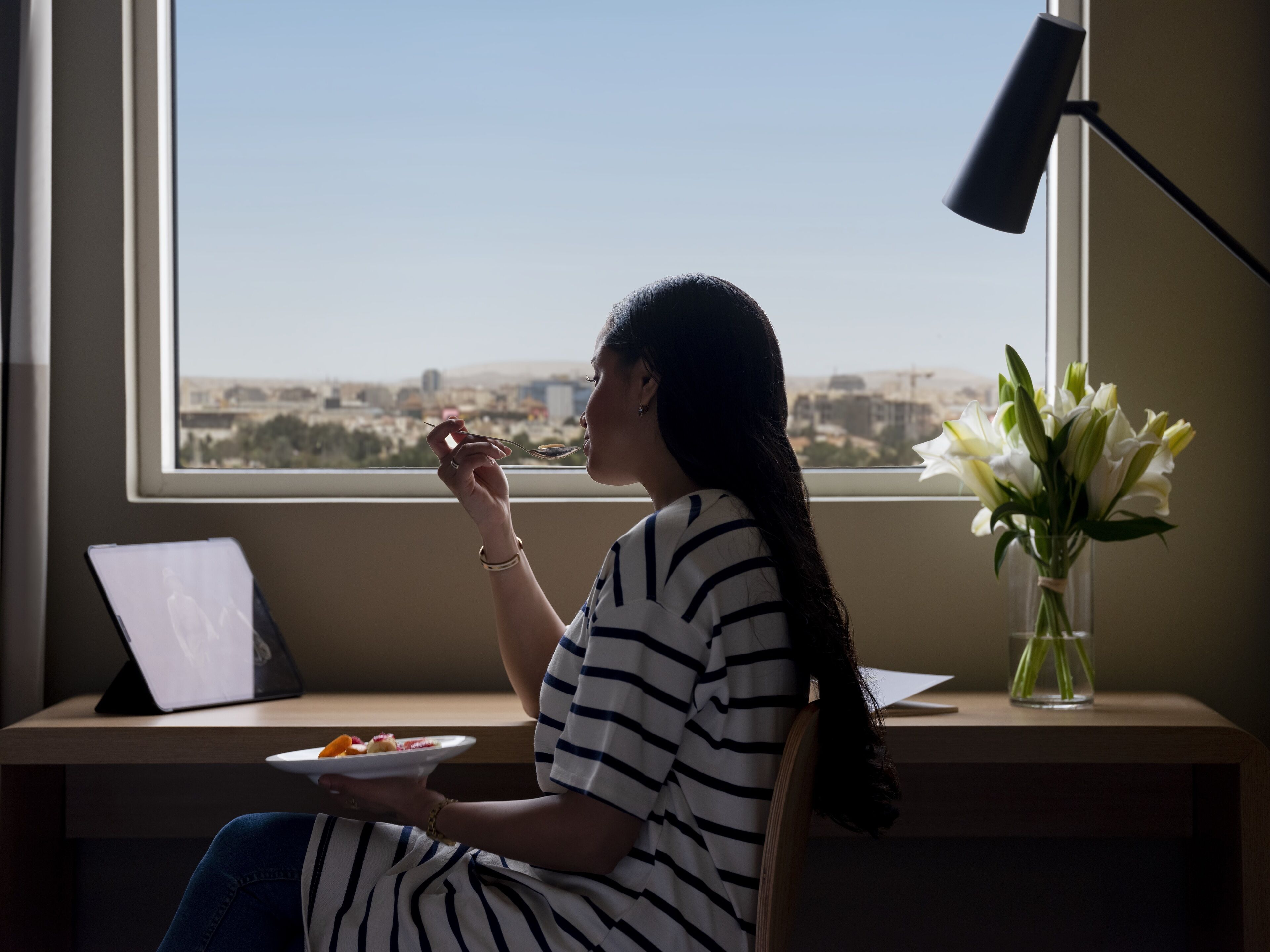 Une personne est assise à un bureau près d’une fenêtre, avec un ordinateur portable, une assiette de nourriture et un vase de fleurs.