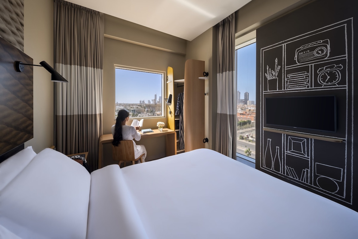 Ein Hotelzimmer mit einem großen Bett, einem Schreibtisch mit Stuhl, einem Fenster mit Blick auf die Stadt und einer Wand mit Kreidezeichnungen.