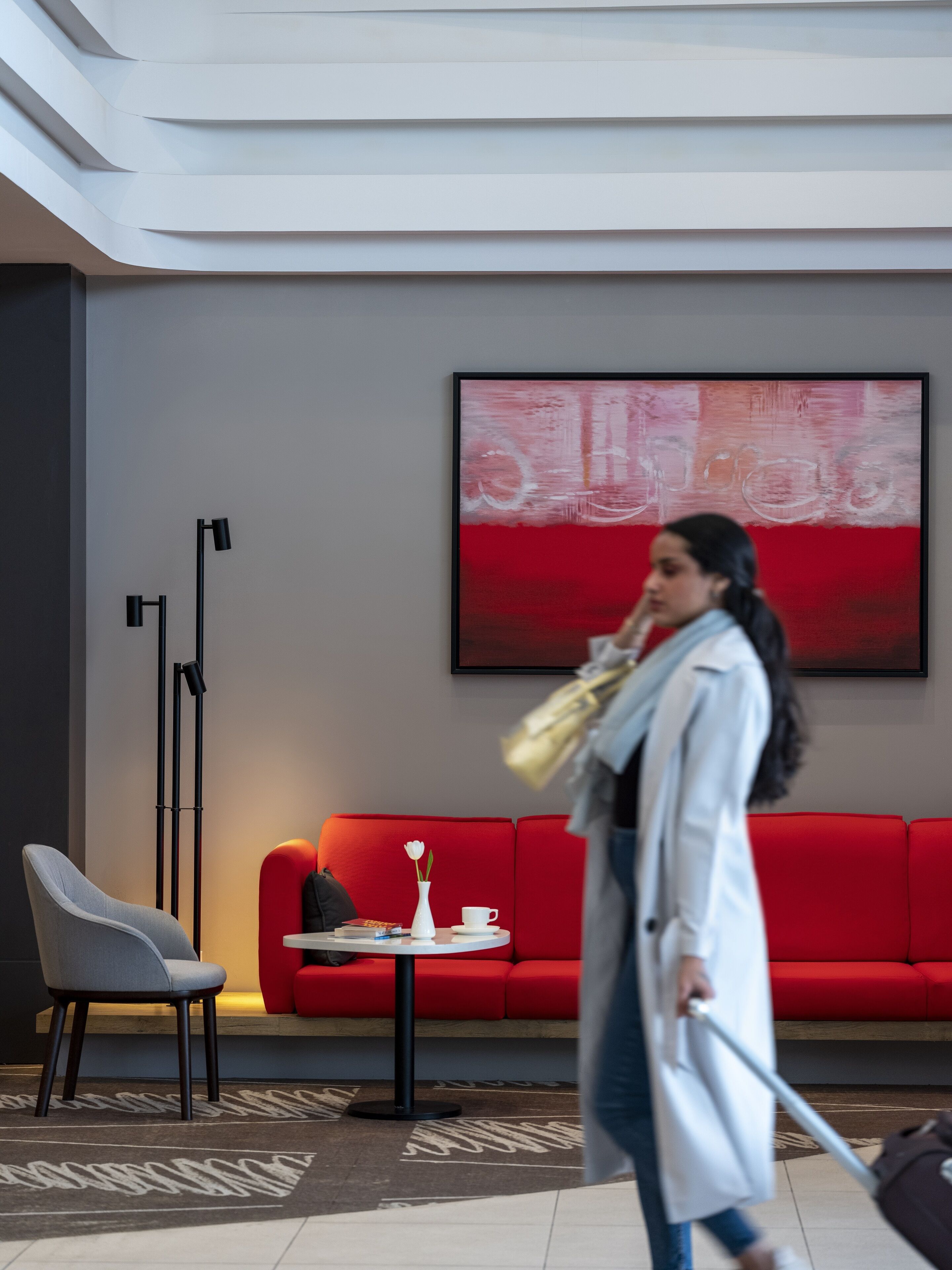 A modern hotel lobby with a red sofa, a white vase with a flower, a black floor lamp, and a large abstract painting on the wall.
