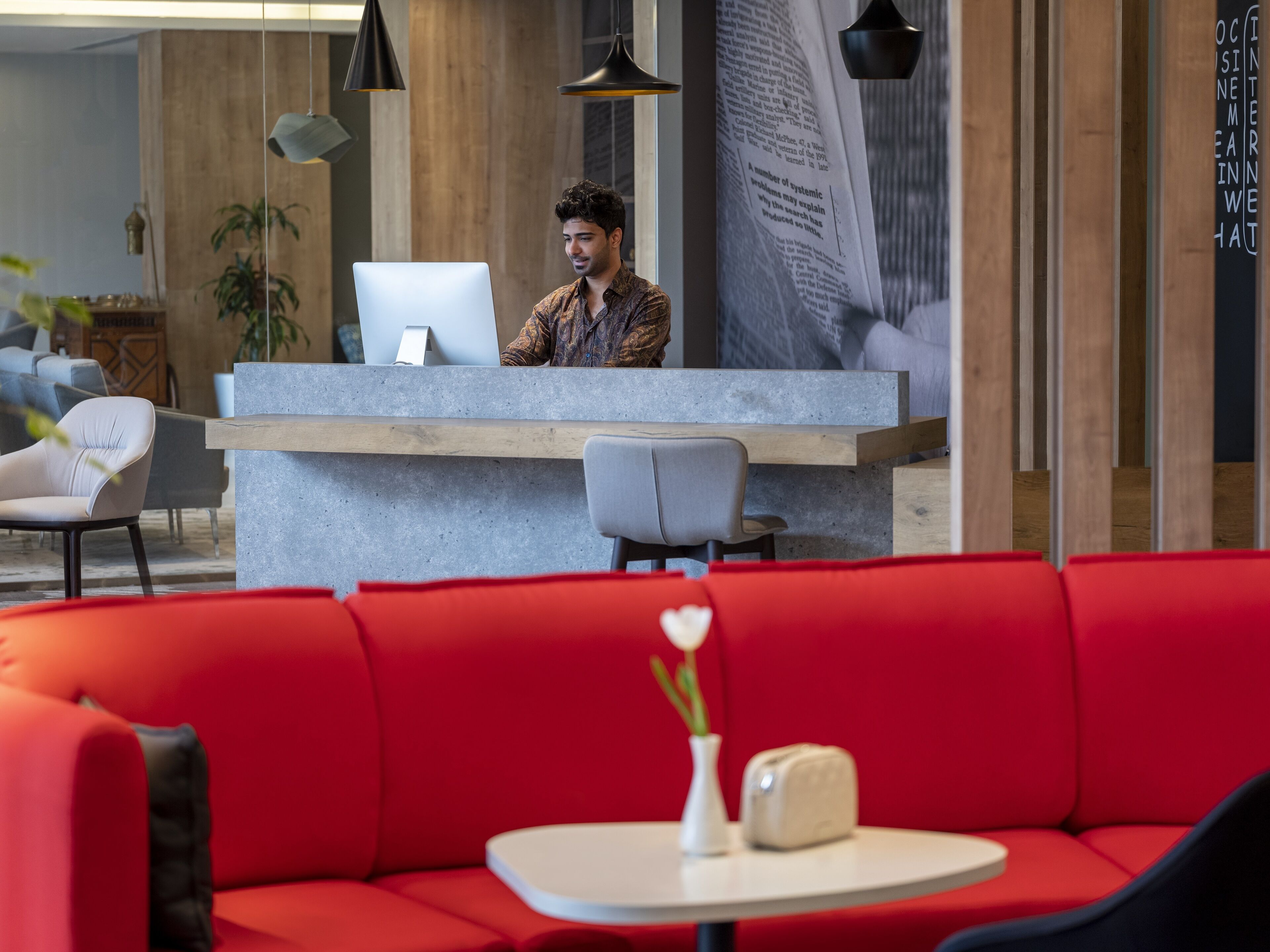 A reception area with a receptionist behind a counter, red seating, and a small table with a vase and a toaster.