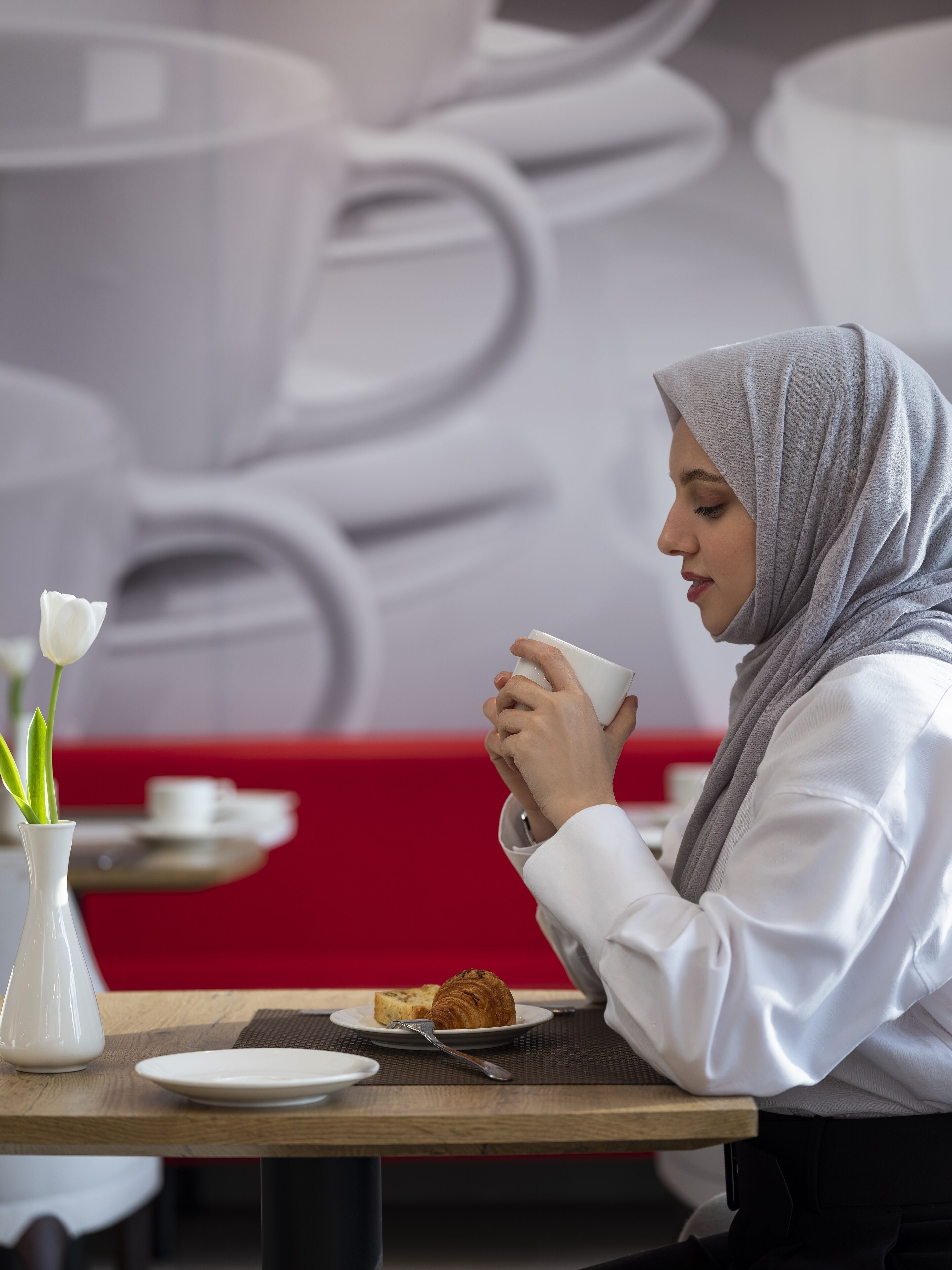 A person seated at a table with a white cup, a plate with a pastry, and a vase with flowers.