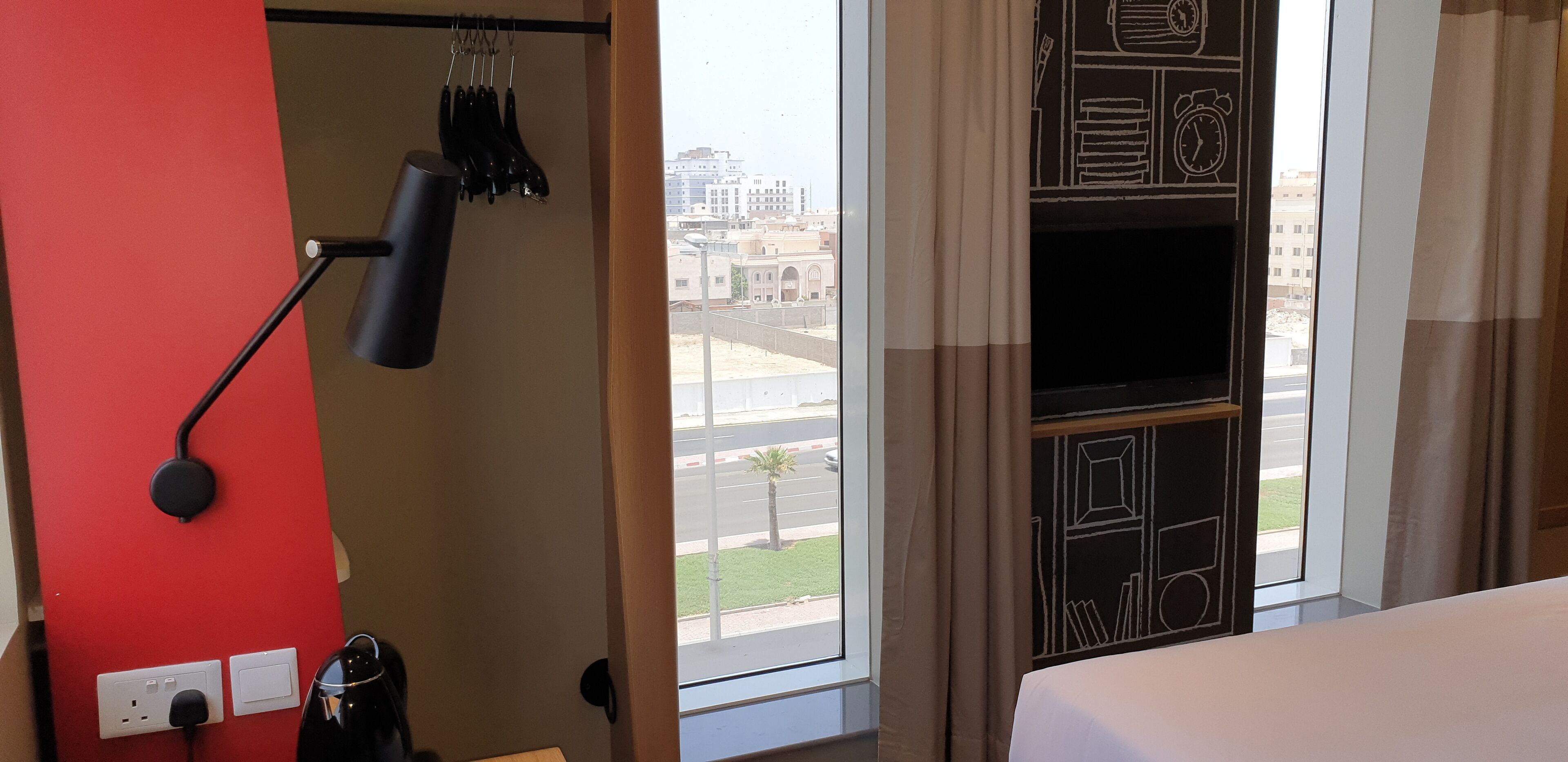 A hotel room with a bed, a red wall, a window with a view of buildings, and a wall-mounted TV.