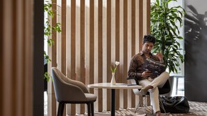 A man sitting at a round table with a white vase, in a room with vertical wooden slats and a large plant.