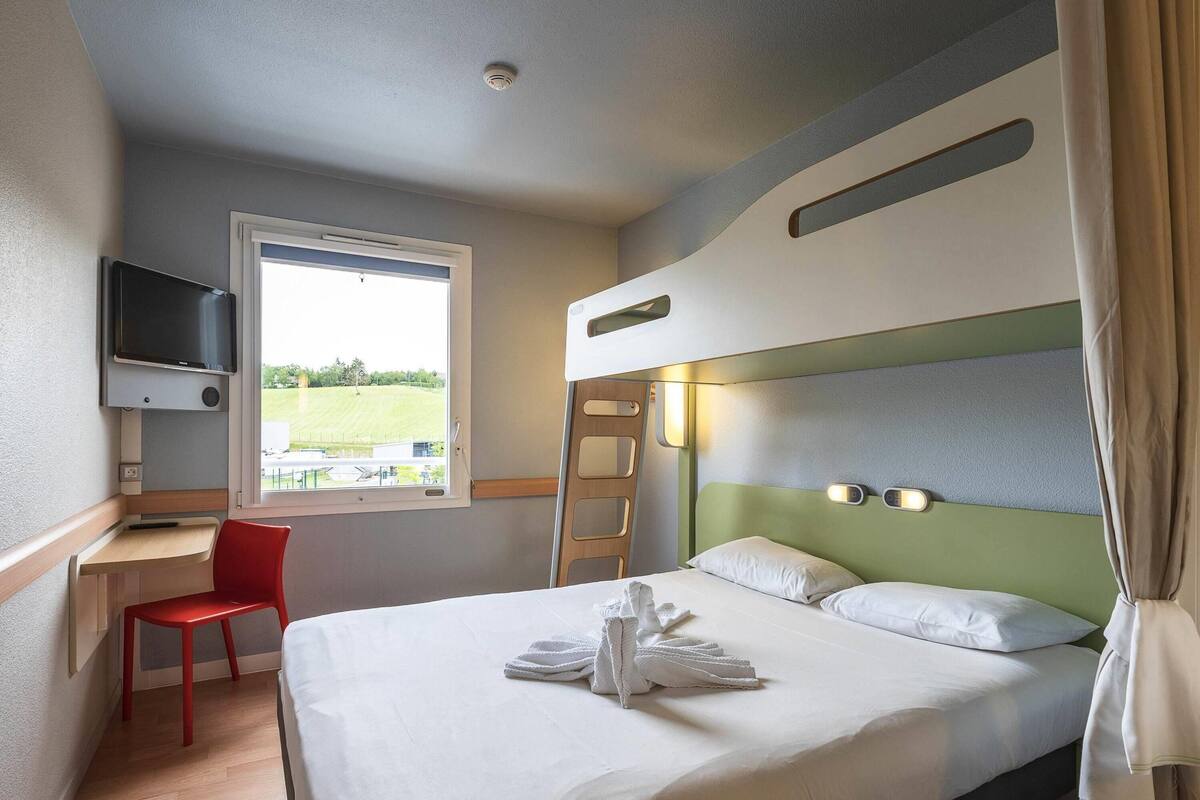 A hotel room with a bed, a desk, a chair, a TV, and a window with a view of a green landscape.