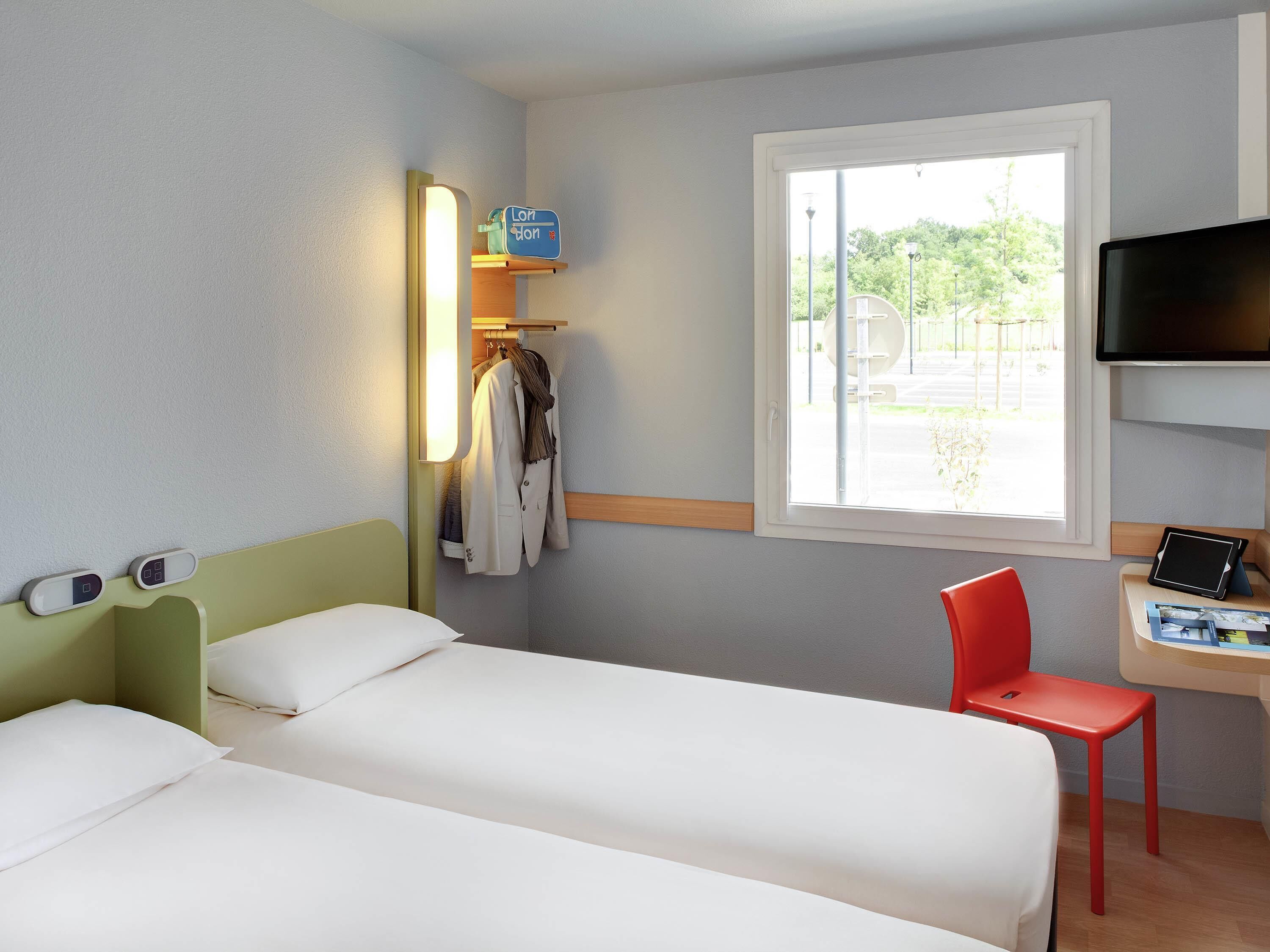 A hotel room with two beds, a desk with a chair, a TV, a window, and a coat rack with clothes.