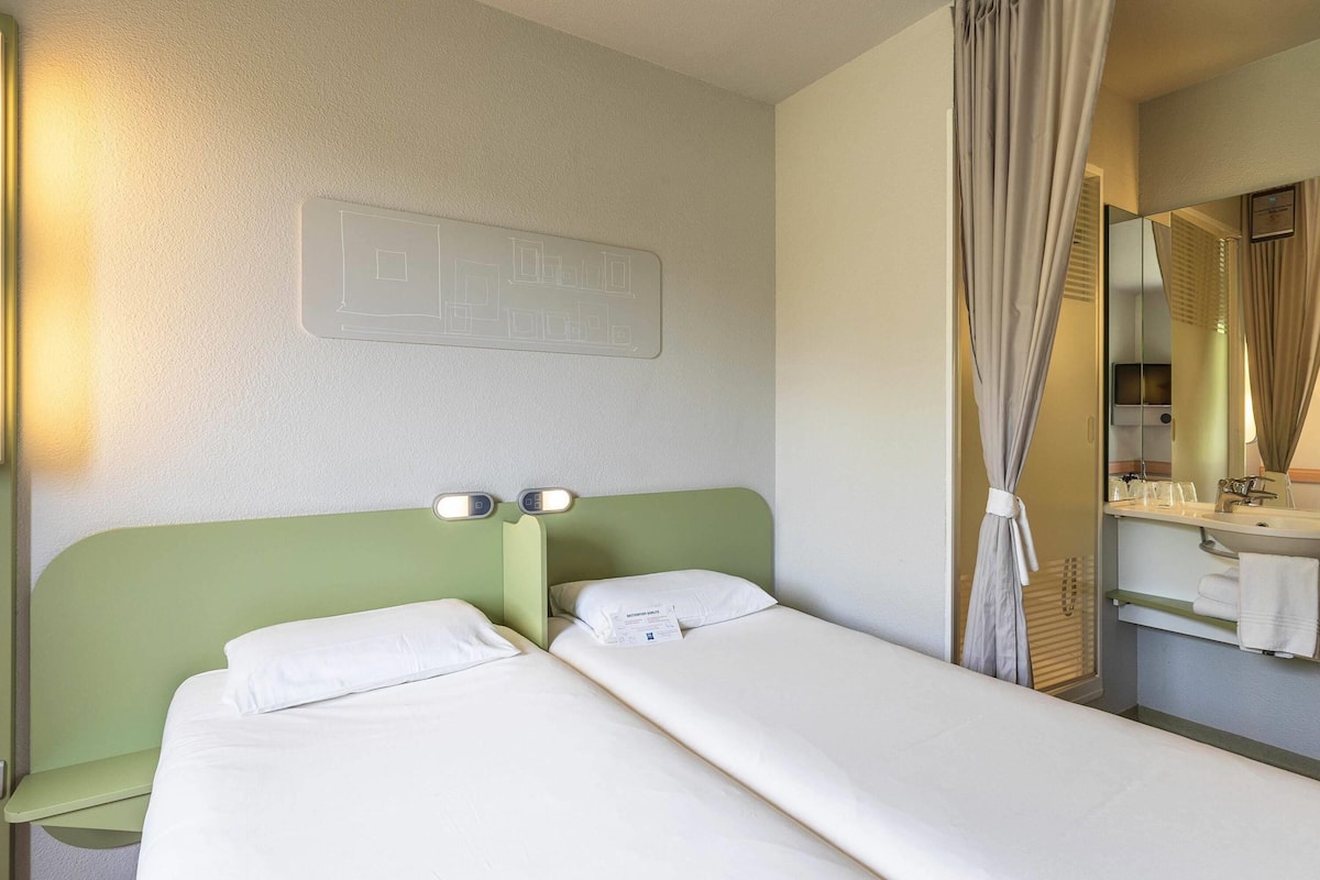 A hotel room with two single beds, a green headboard, a wall-mounted light panel, a bathroom with a mirror, and a curtain.