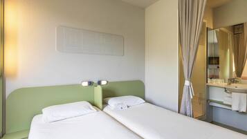 A hotel room with two single beds, a green headboard, a wall-mounted light panel, a bathroom with a mirror, and a curtain.