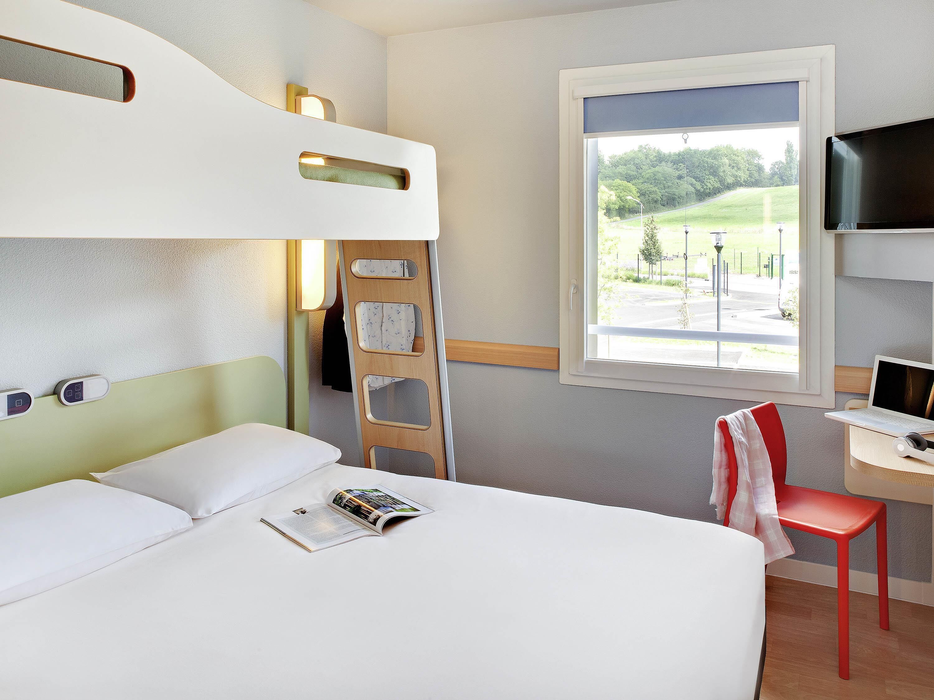 A hotel room with a bunk bed, a desk with a laptop, a chair, and a window with a view of greenery.