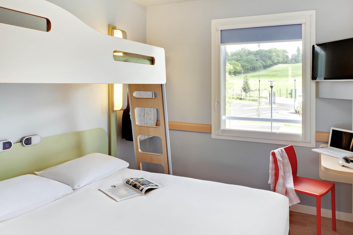A hotel room with a bunk bed, a desk with a laptop, a chair, and a window with a view of greenery.
