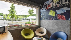 A lounge area with a view of a parking lot, a wall with a bookshelf mural, and three chairs. - ibis budget Saint-Pourçain-sur-Sioule (Saint-Pourcain-sur-Sioule)