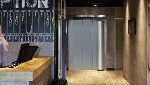 A reception area with a wooden counter, a chalkboard with text, and a modern elevator. - ibis budget Saint-Pourçain-sur-Sioule (Saint-Pourcain-sur-Sioule)
