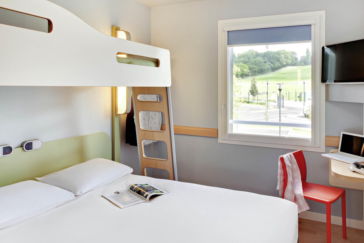 A hotel room with a bunk bed, a desk with a laptop, a chair, and a window with a view of greenery.