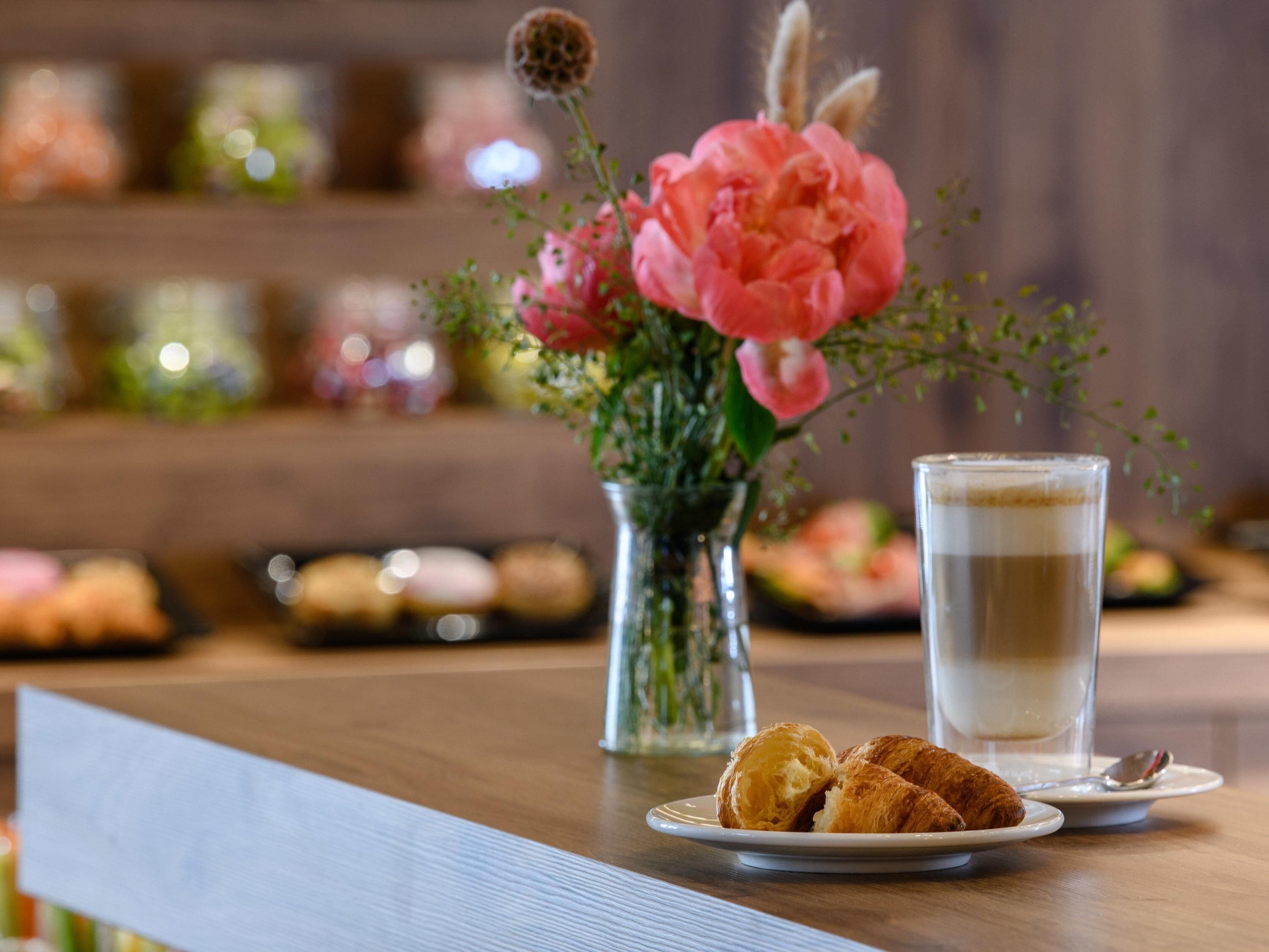 A table with a plate of croissants, a glass of coffee, and a vase with pink flowers.