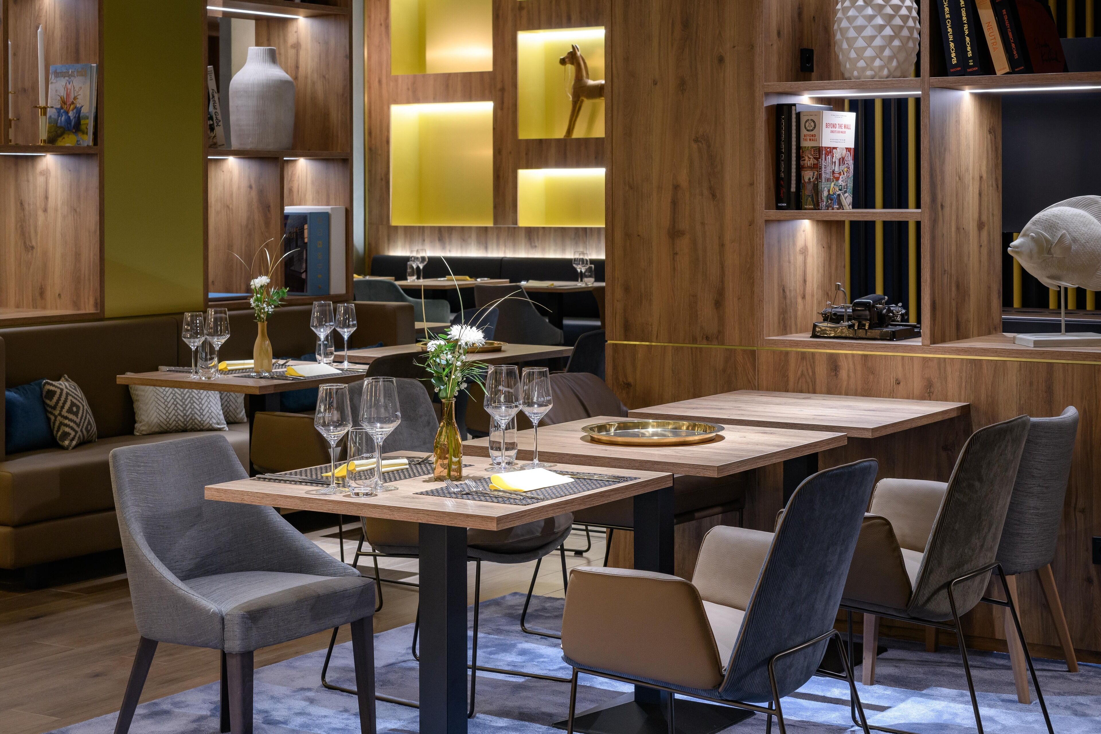 A modern restaurant interior with wooden paneling, tables set with wine glasses, and a bookshelf with books and decorative items.