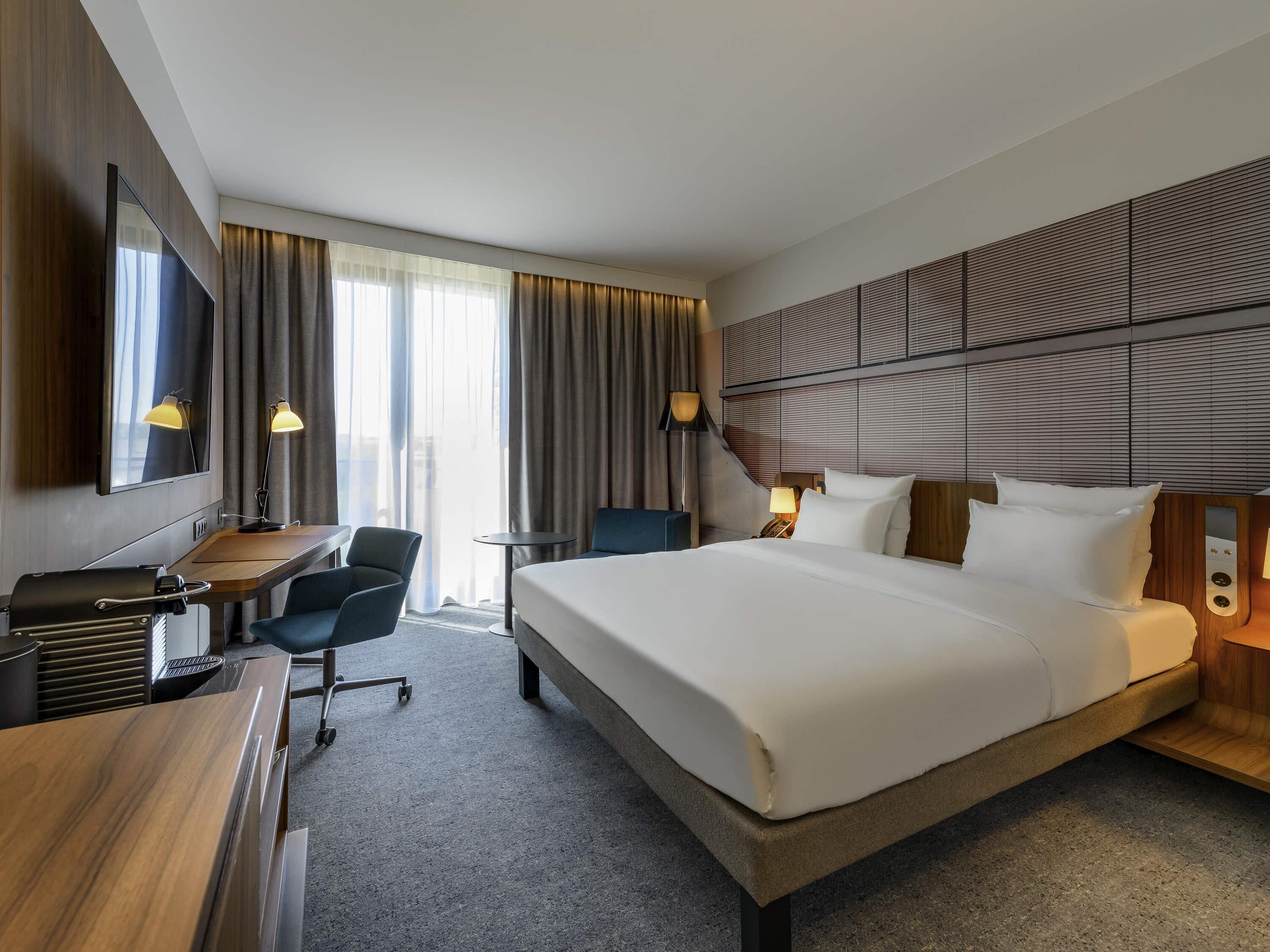 A hotel room with a large bed, a desk, a chair, a TV, and a lamp.