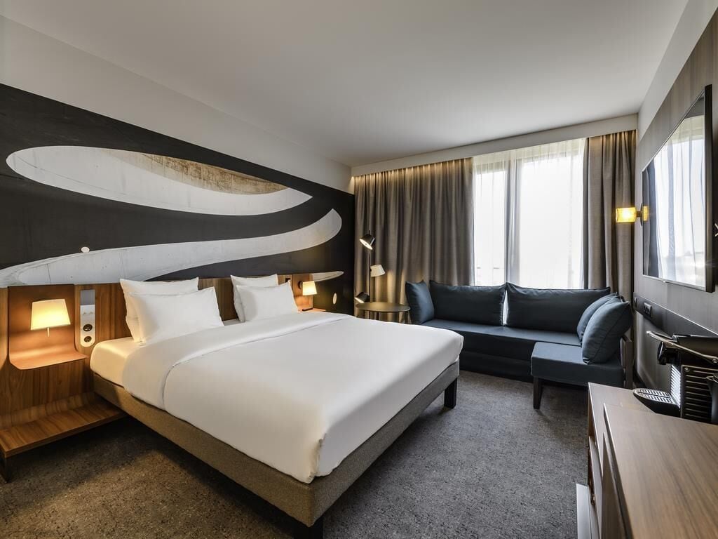 A modern hotel room with a large bed, a sofa, a desk, and a TV.