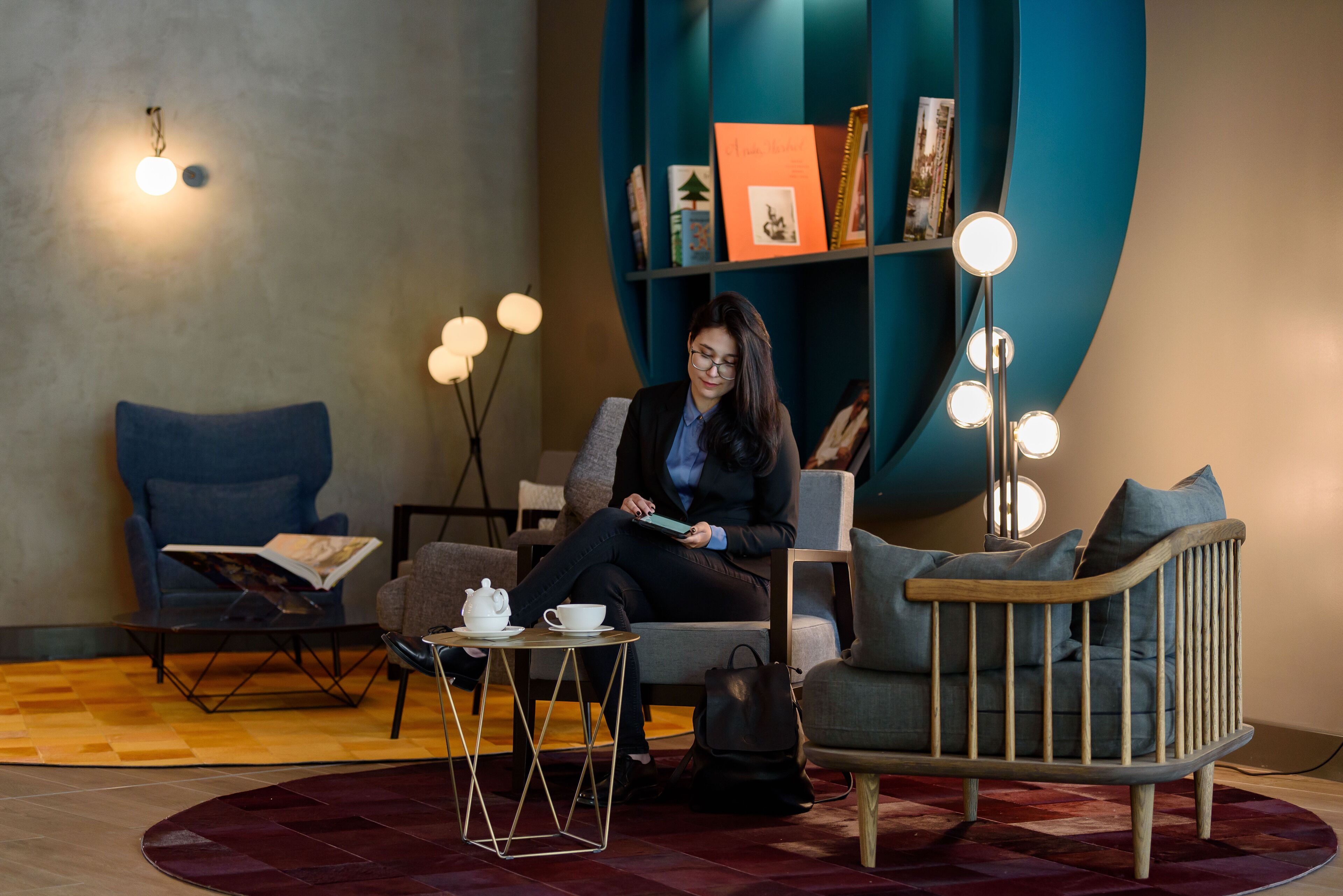 Een moderne zithoek met een bank, een kleine tafel met theepot en kopje, een boekenkast met boeken en decoratieve objecten, en een vloerlamp.