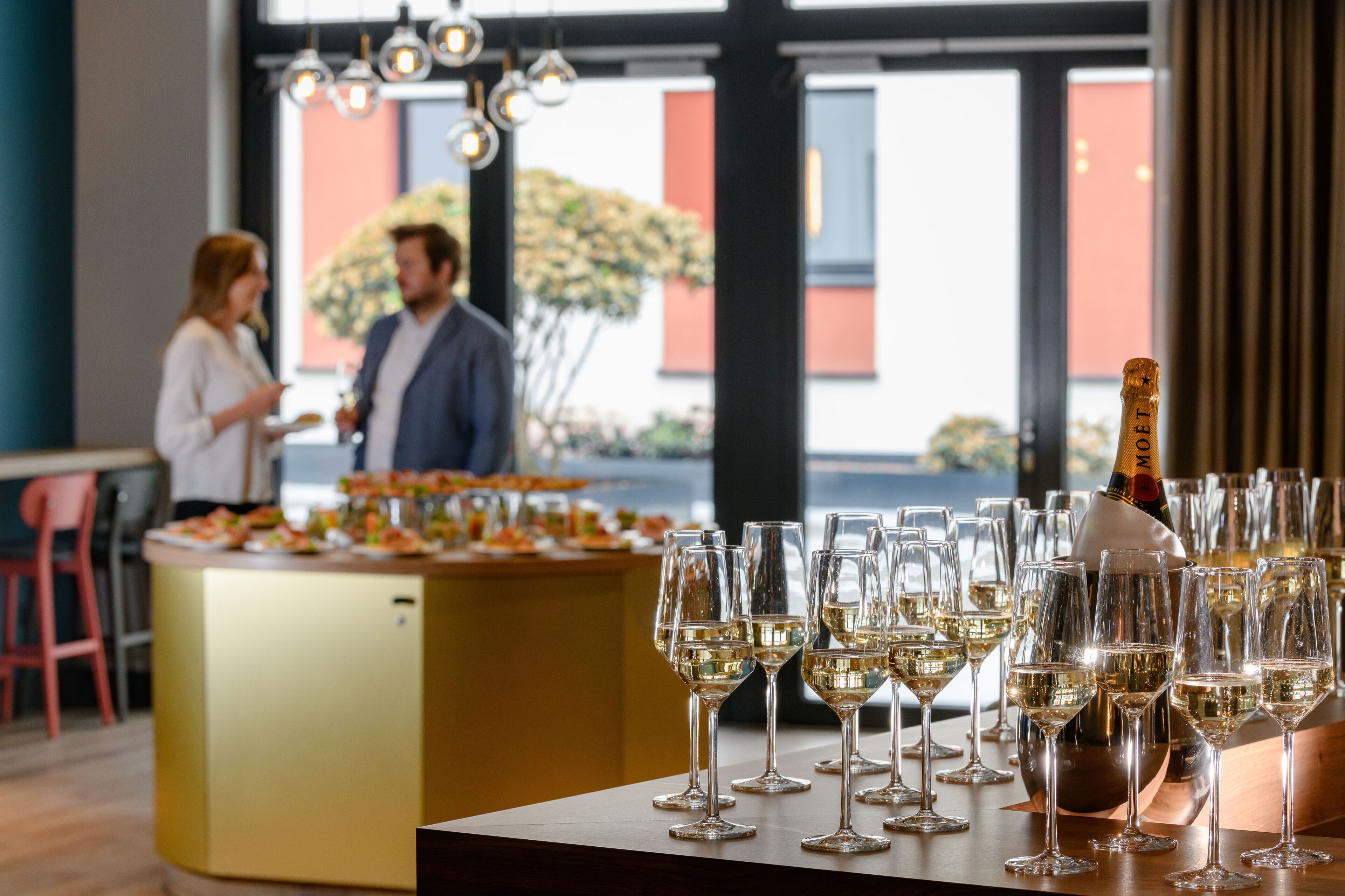 A bar area with multiple glasses of white wine and a bottle of champagne. There is a bar counter with a gold finish and a woman and a man in the background.