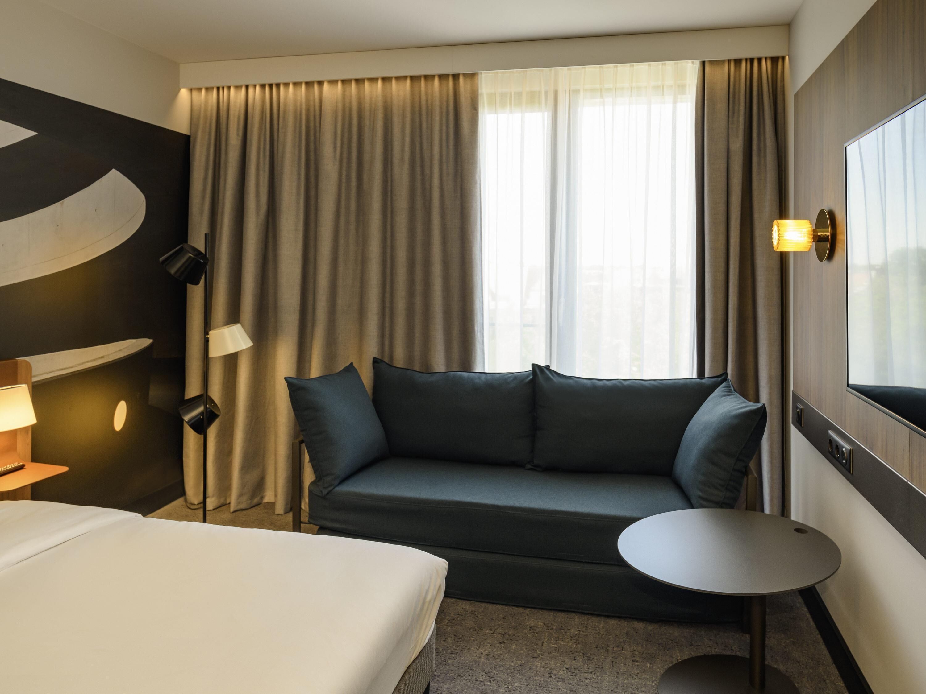 Una habitación de hotel con cama, sofá, una mesita y un ventanal con cortinas.