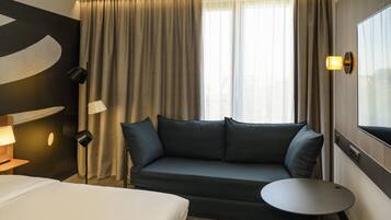 Una habitación de hotel con cama, sofá, una mesita y un ventanal con cortinas.