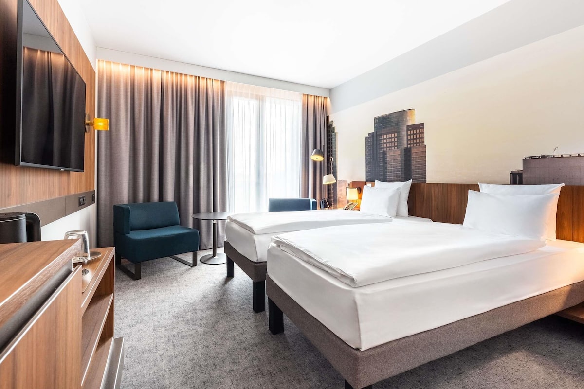 Habitación de hotel con una cama grande, dos sillones, una mesita, televisor montado en la pared y vistas a la ciudad.