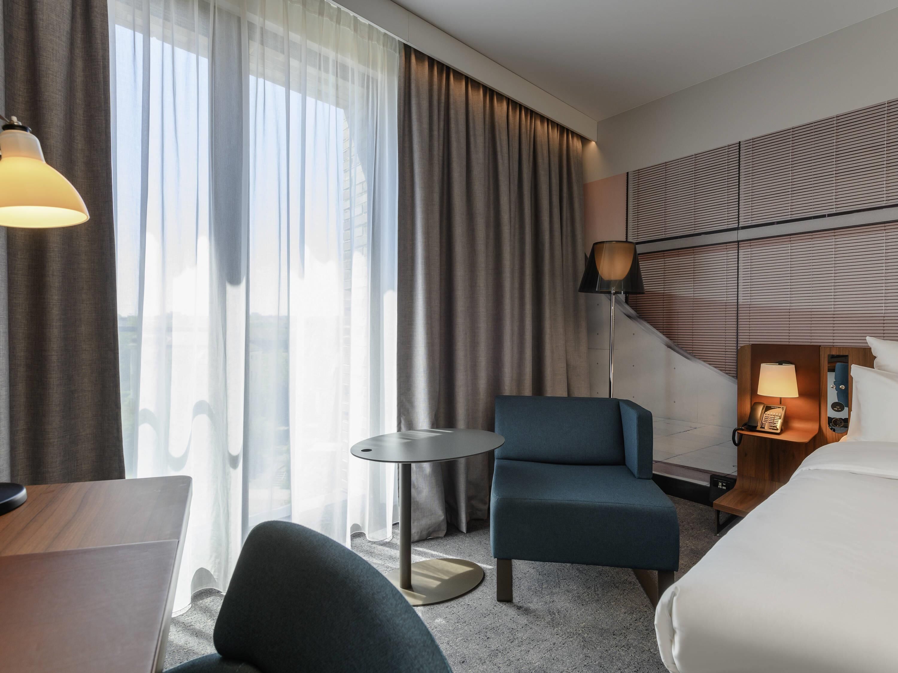 A hotel room with a bed, a desk, a chair, a small table, a lamp, and a view of the outside through large windows.