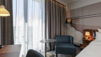 A hotel room with a bed, a desk, a chair, a small table, a lamp, and a view of the outside through large windows.