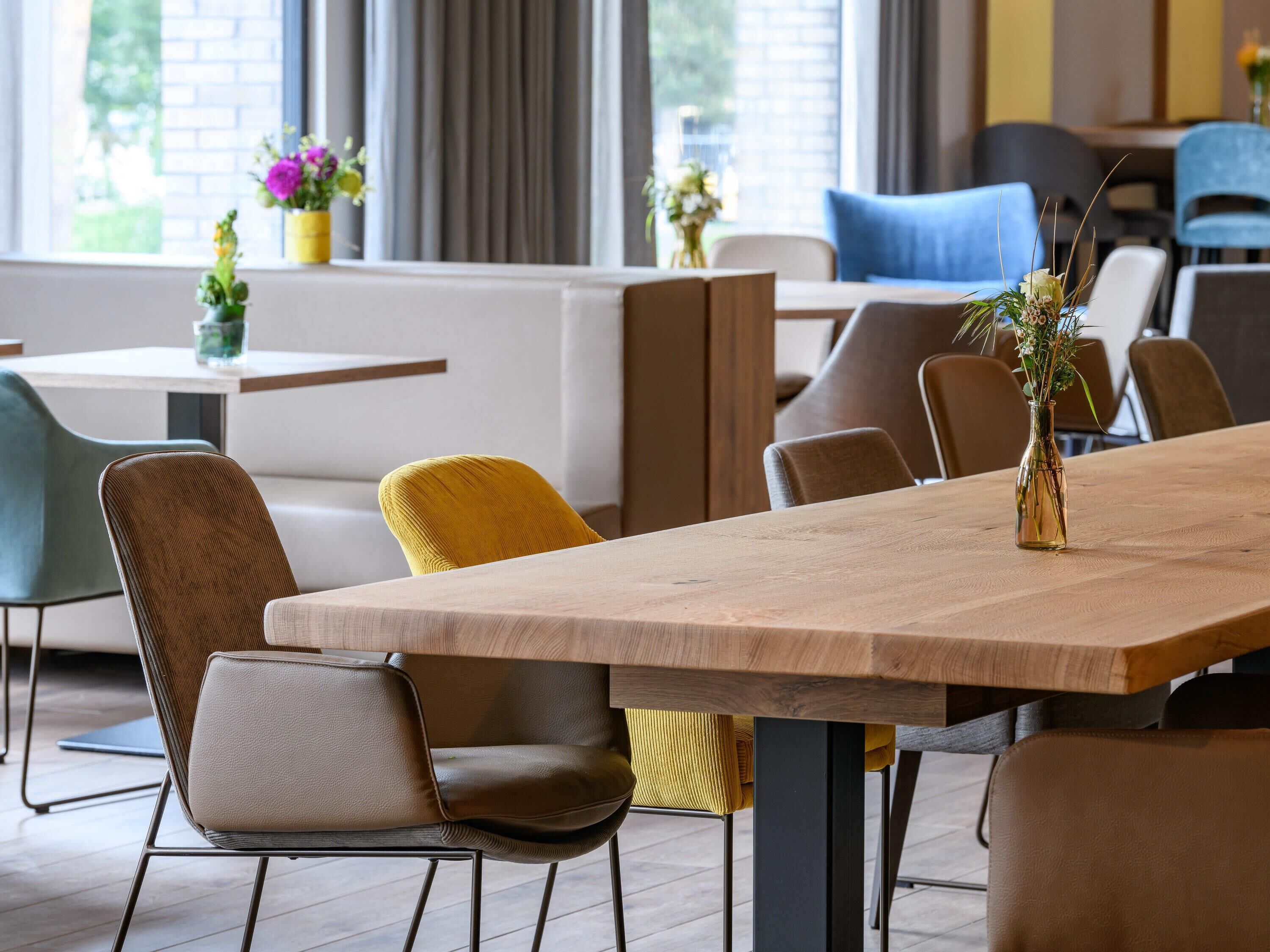 A modern dining area with wooden tables and chairs, a vase with flowers, and a view of the outdoors through large windows.