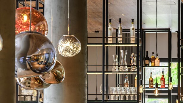Zona de bar moderna con lámparas colgantes de cristal, estantes con botellas y vasos, y una vista a través de una ventana.