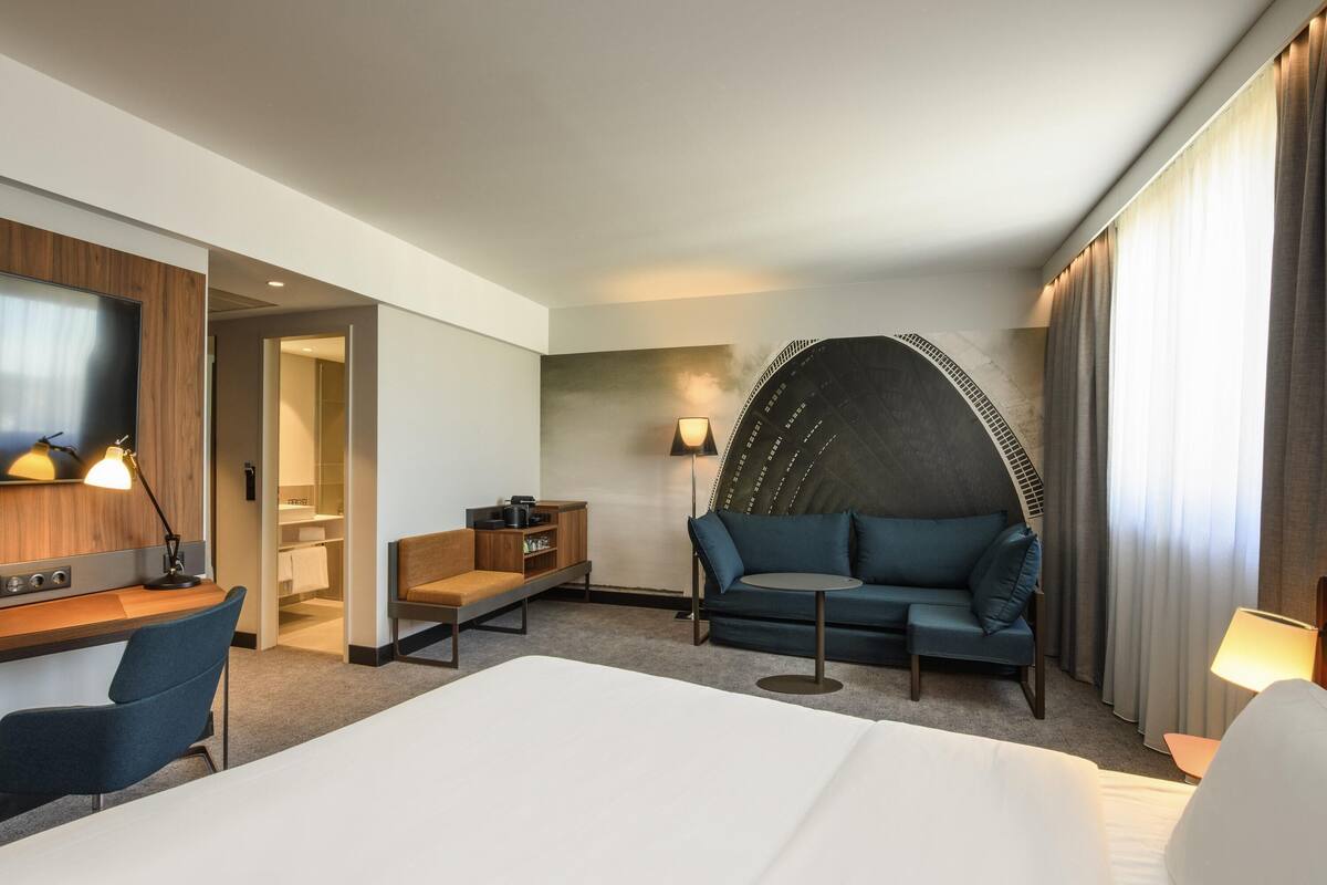 Una habitación de hotel con una cama, un escritorio con una silla, un sofá, un televisor y una lámpara.