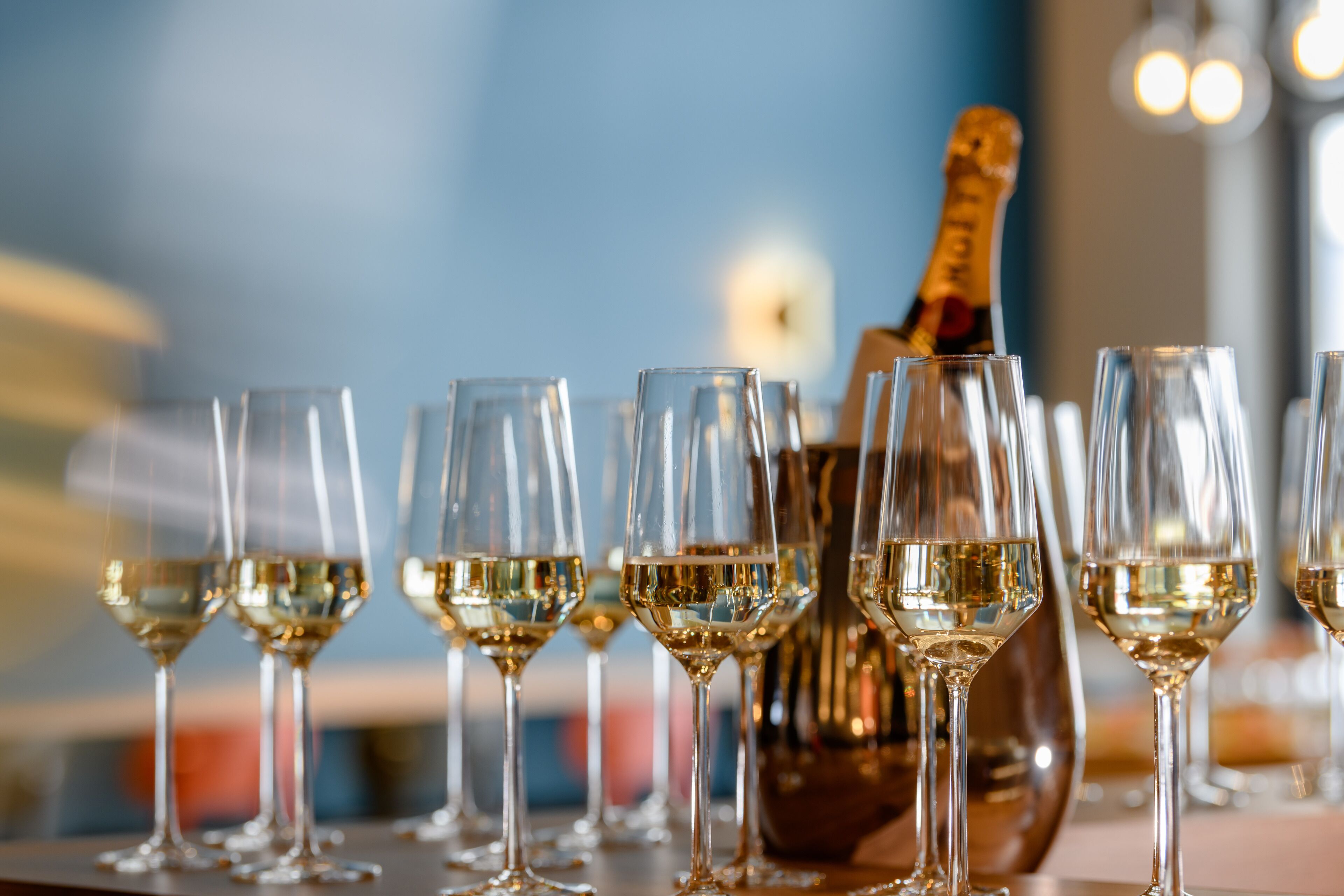 A row of empty wine glasses on a bar counter with a bottle of champagne in the background.
