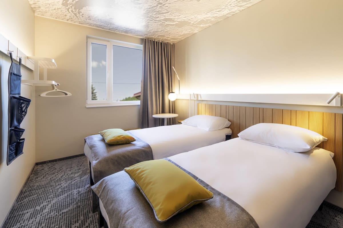 Habitación de hotel con dos camas, una ventana, una lámpara y un perchero de pared.