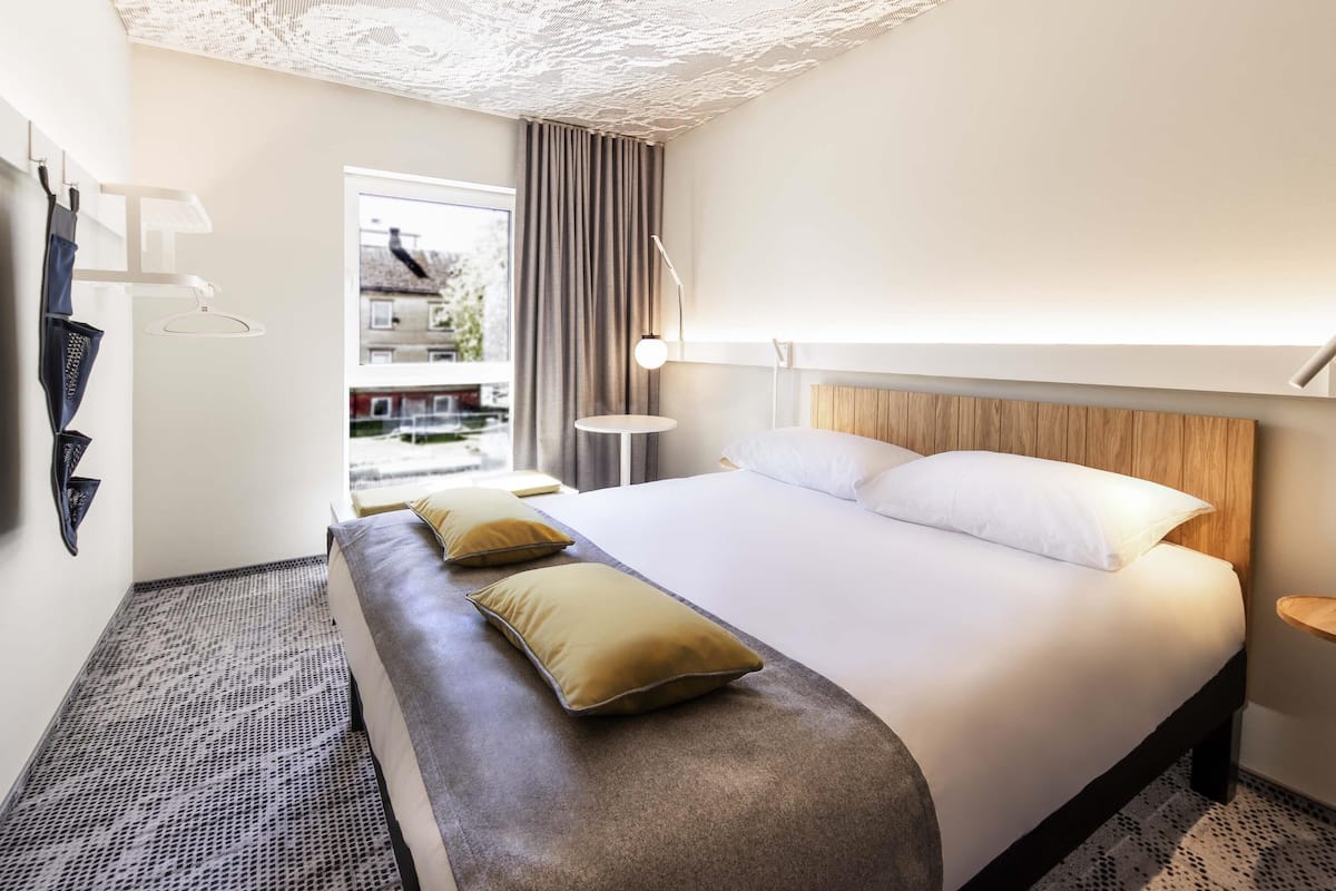 Habitación de hotel con una cama grande, un banco, una mesita y vistas al exterior.