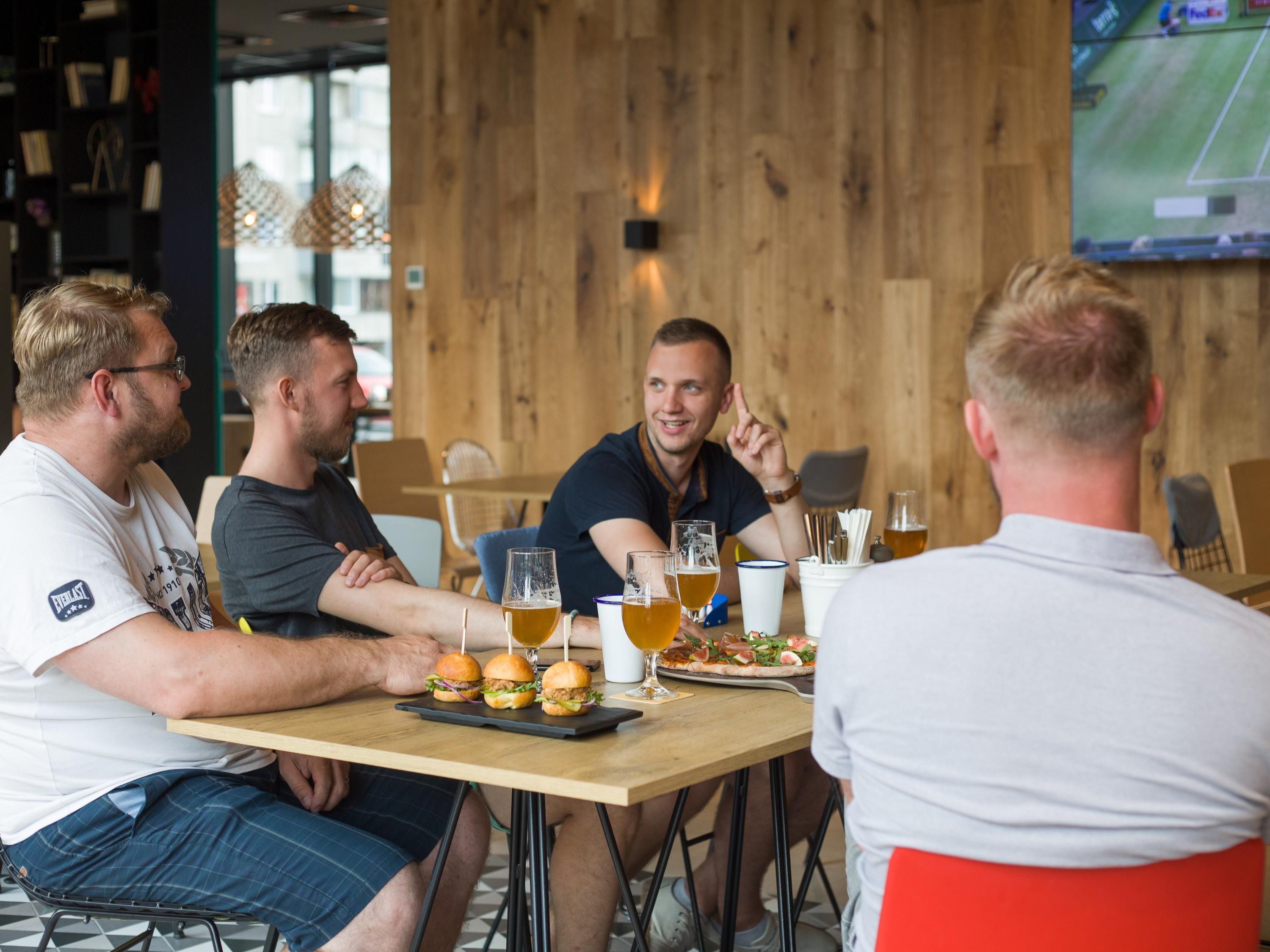 A group of people seated at a wooden table with glasses of beer and food, in a room with a wooden wall and a TV screen displaying a sports game.