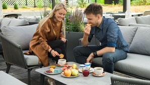Ein Paar sitzt auf einem grauen Sofa, wobei ein Tisch mit Frühstücksleckereien wie Eiern, Toast und Obst zwischen ihnen steht.
