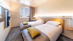 Ein Hotelzimmer mit zwei Betten, einem Fenster, einem an der Wand montierten Fernseher und einem kleinen Nachttisch.