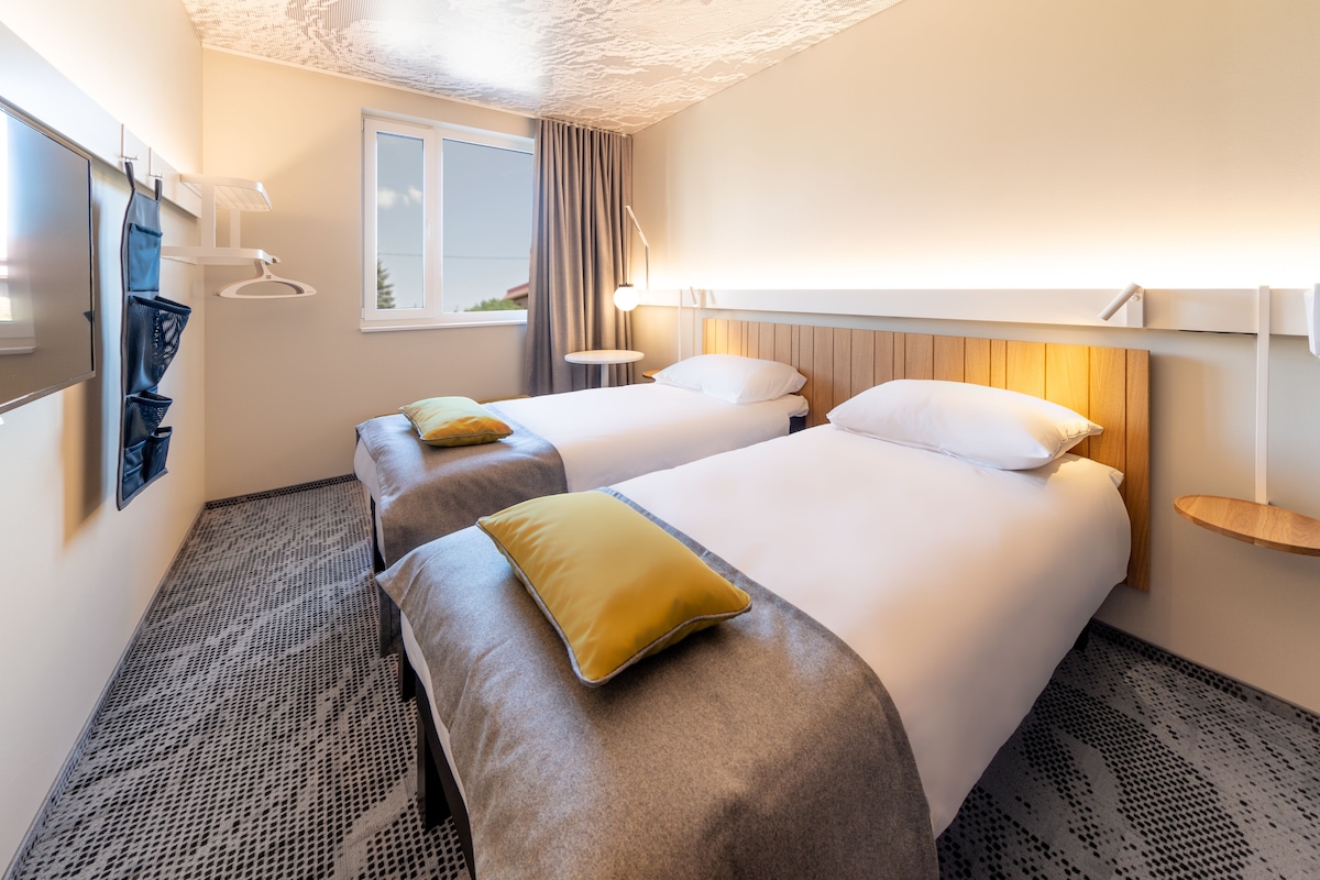 Une chambre d’hôtel avec deux lits, une fenêtre, un téléviseur fixé au mur et une petite table de chevet.