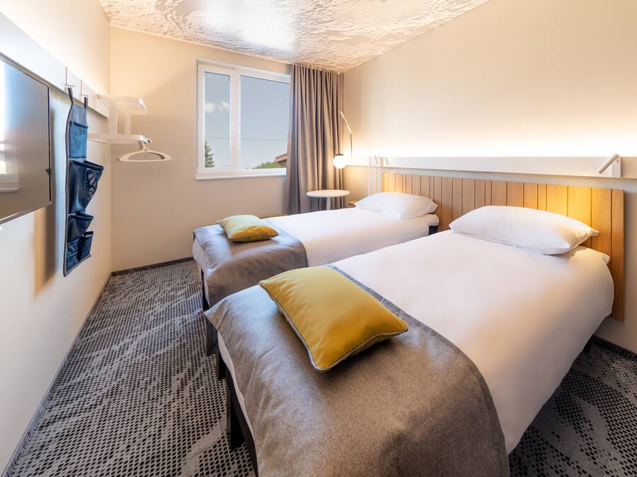 Ein Hotelzimmer mit zwei Betten, einem Fenster, einem an der Wand montierten Fernseher und einem kleinen Nachttisch.