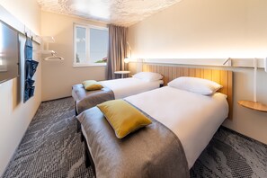 Ein Hotelzimmer mit zwei Betten, einem Fenster, einem an der Wand montierten Fernseher und einem kleinen Nachttisch.