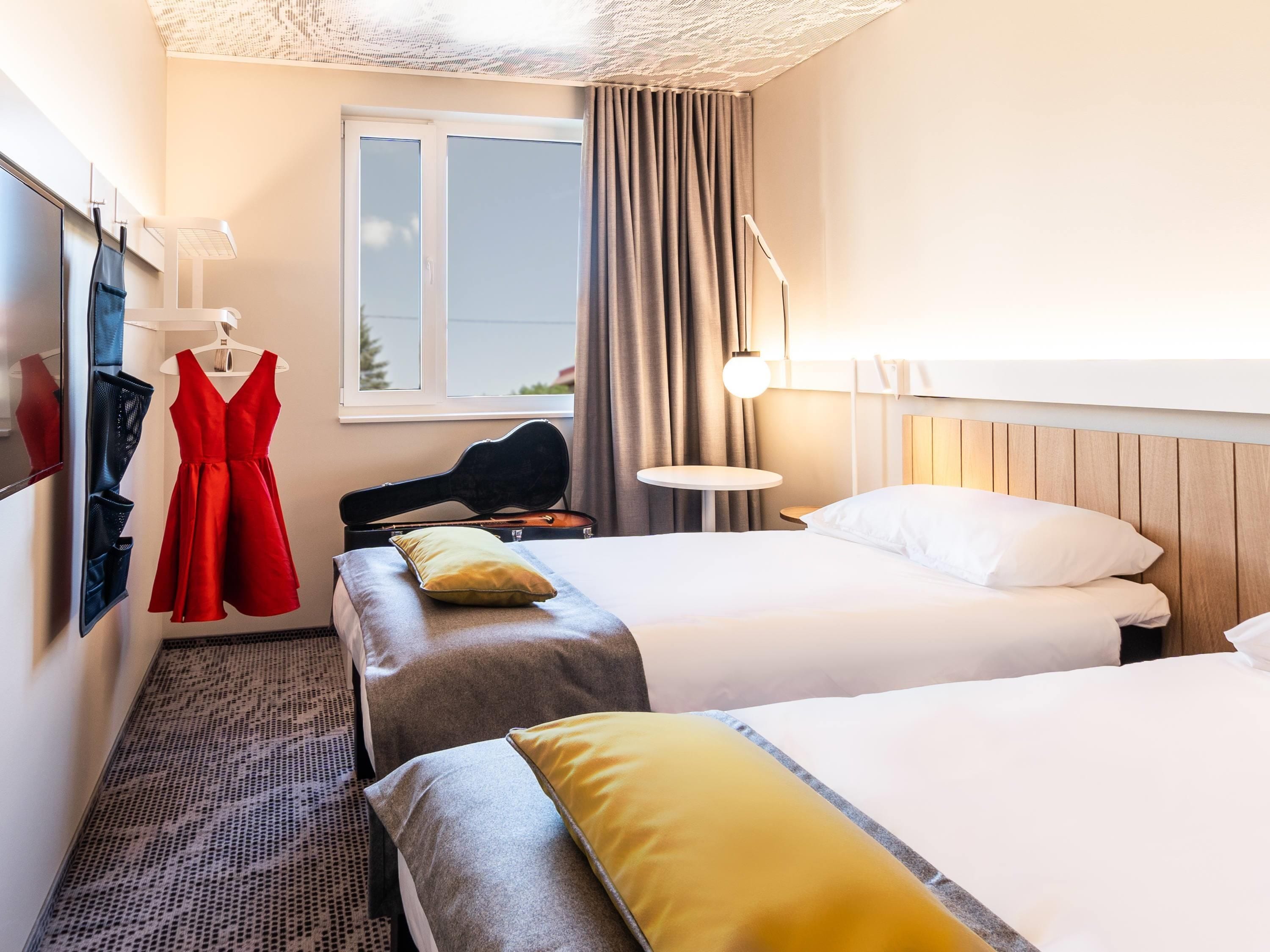 Habitación de hotel con dos camas, un vestido rojo colgado en la pared, una guitarra y una ventana con cortinas.