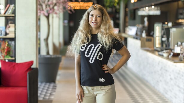 Eine Frau in einem schwarzen T-Shirt mit den Aufschriften 'GO' und 'ibis' steht in einem modernen Innenraum mit einem roten Sofa, Bücherregalen und einer Theke mit Gegenständen.