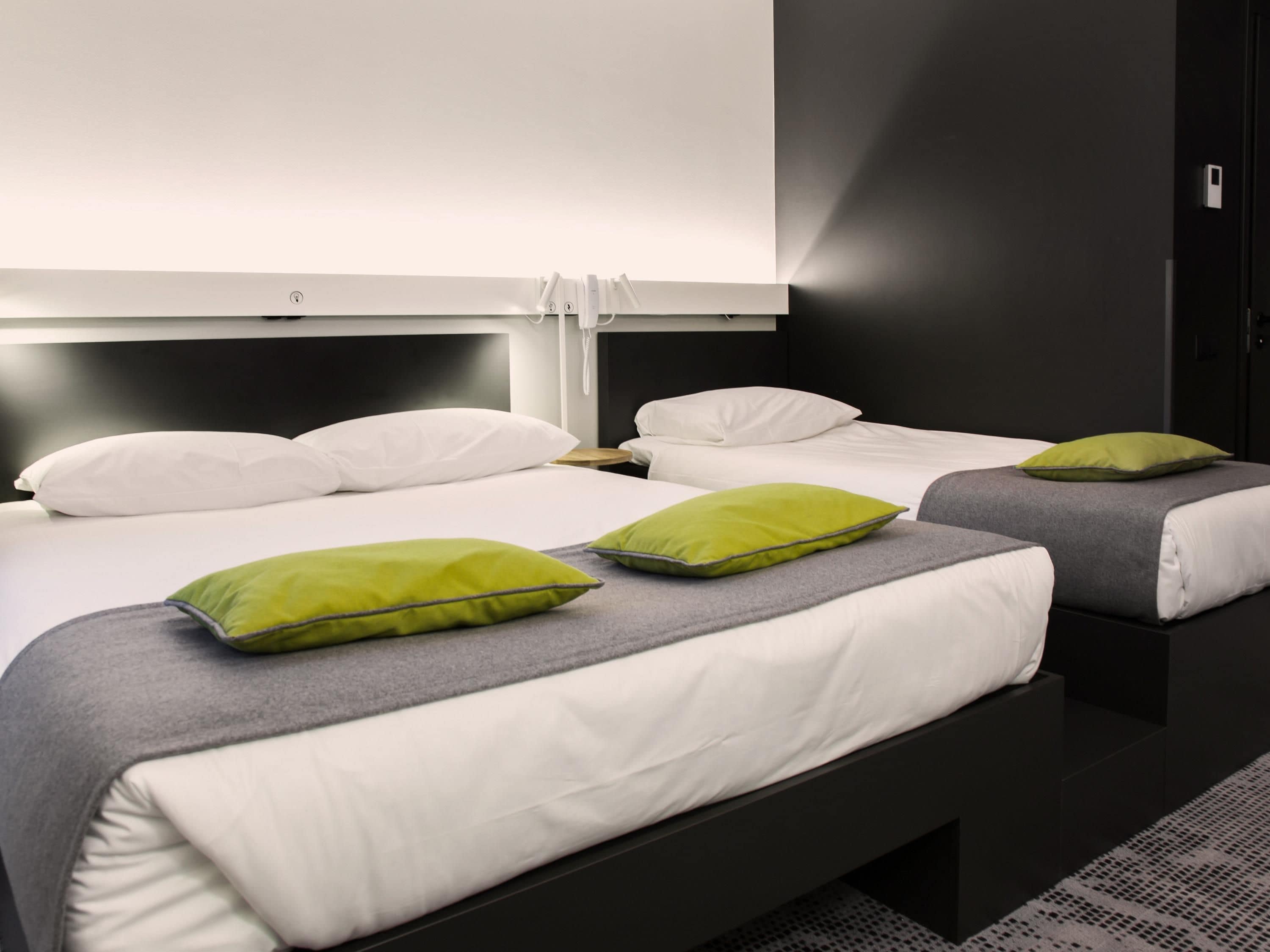 Habitación de hotel con dos camas, cada una con ropa de cama blanca y almohadas verdes, un televisor de pantalla plana grande montado en la pared y un armario de color oscuro.