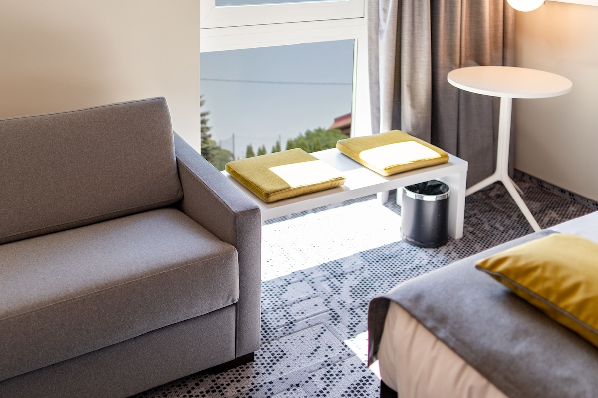 Une chambre d’hôtel moderne avec un canapé gris, une petite table blanche sur laquelle se trouve un plateau avec des articles de toilette, une petite poubelle et un lit avec un oreiller jaune.