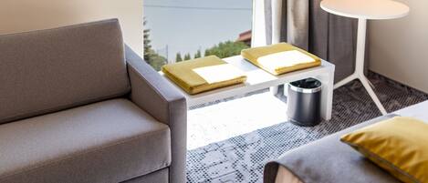 A modern hotel room with a grey sofa, a small white table with a tray of toiletries, a compact trash bin, and a bed with a yellow pillow.